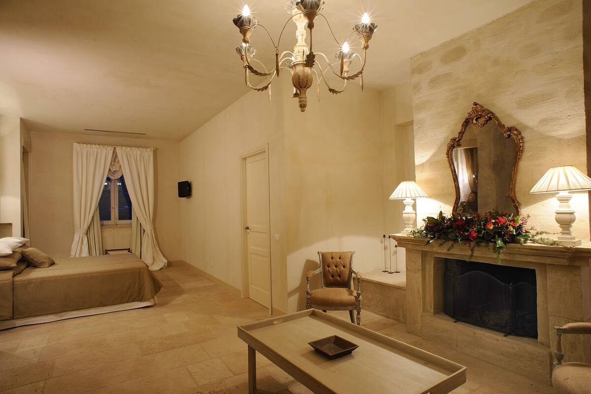 Quarto de hotel com cama, poltrona, mesa, lareira e lustre.