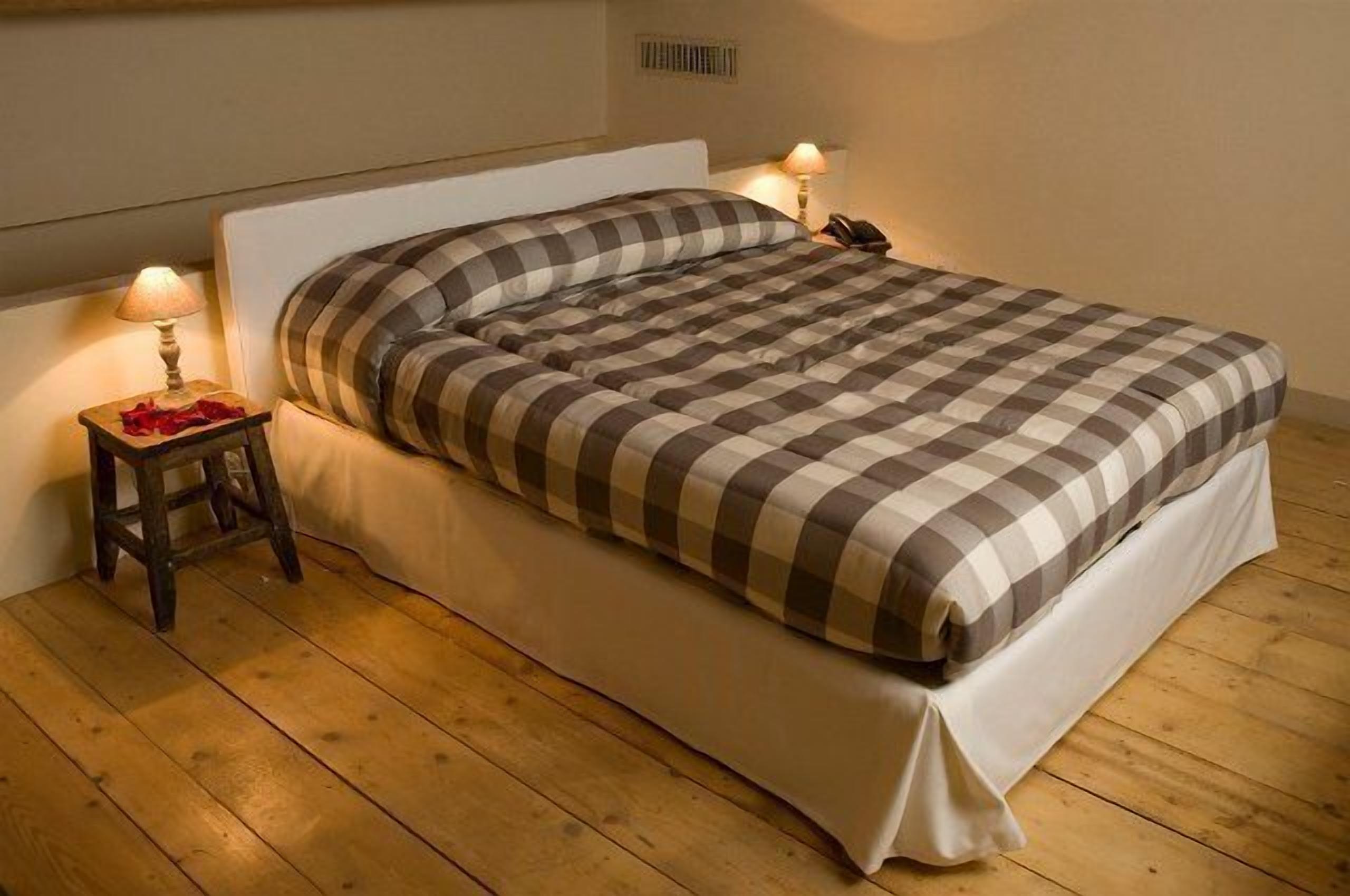 Un lit recouvert d’une couverture à carreaux, une table de chevet avec une lampe et un petit tabouret sur lequel se trouve un objet rouge.