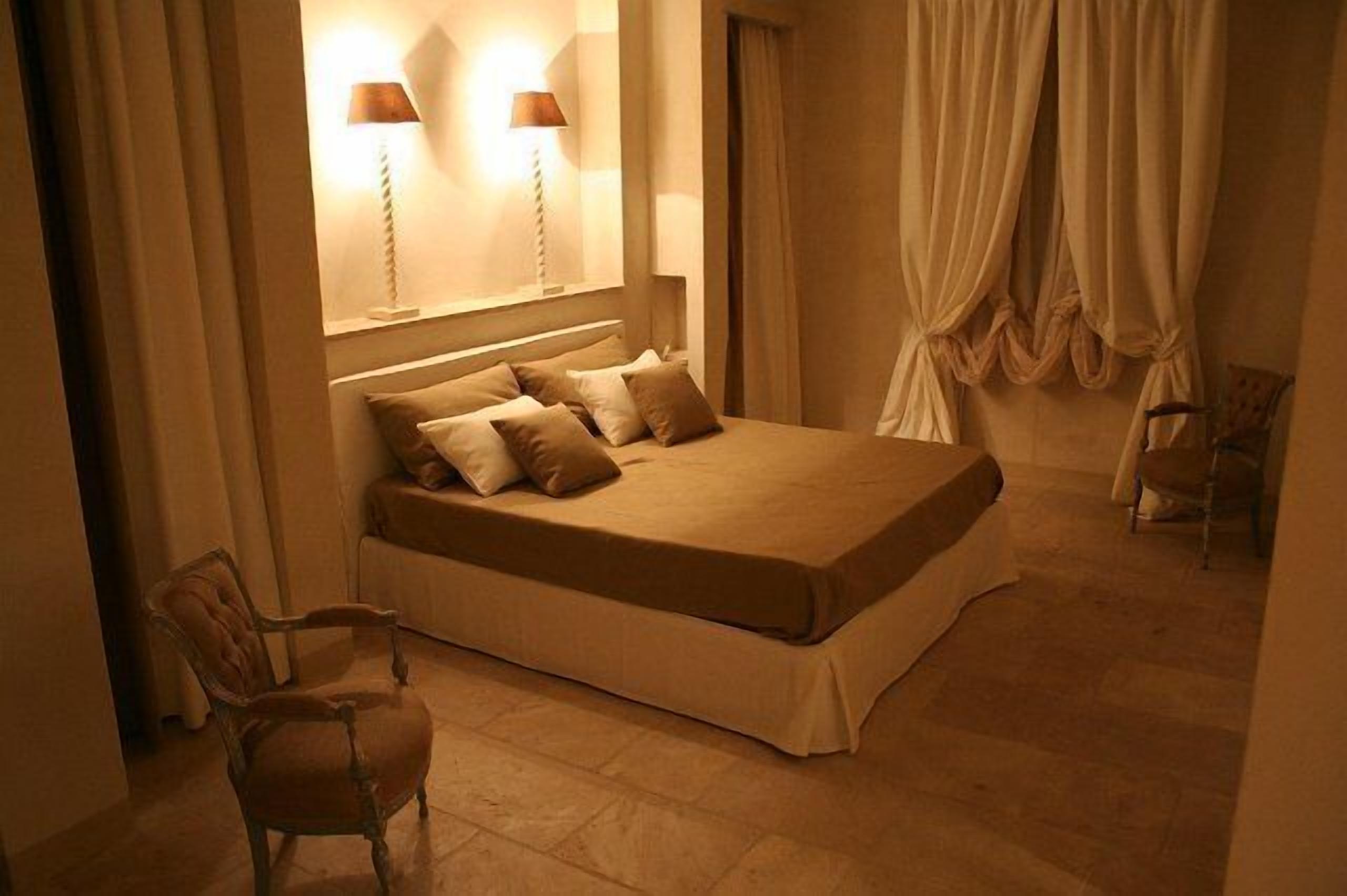 A bedroom with a large bed, two bedside lamps, a chair, and a window with curtains.