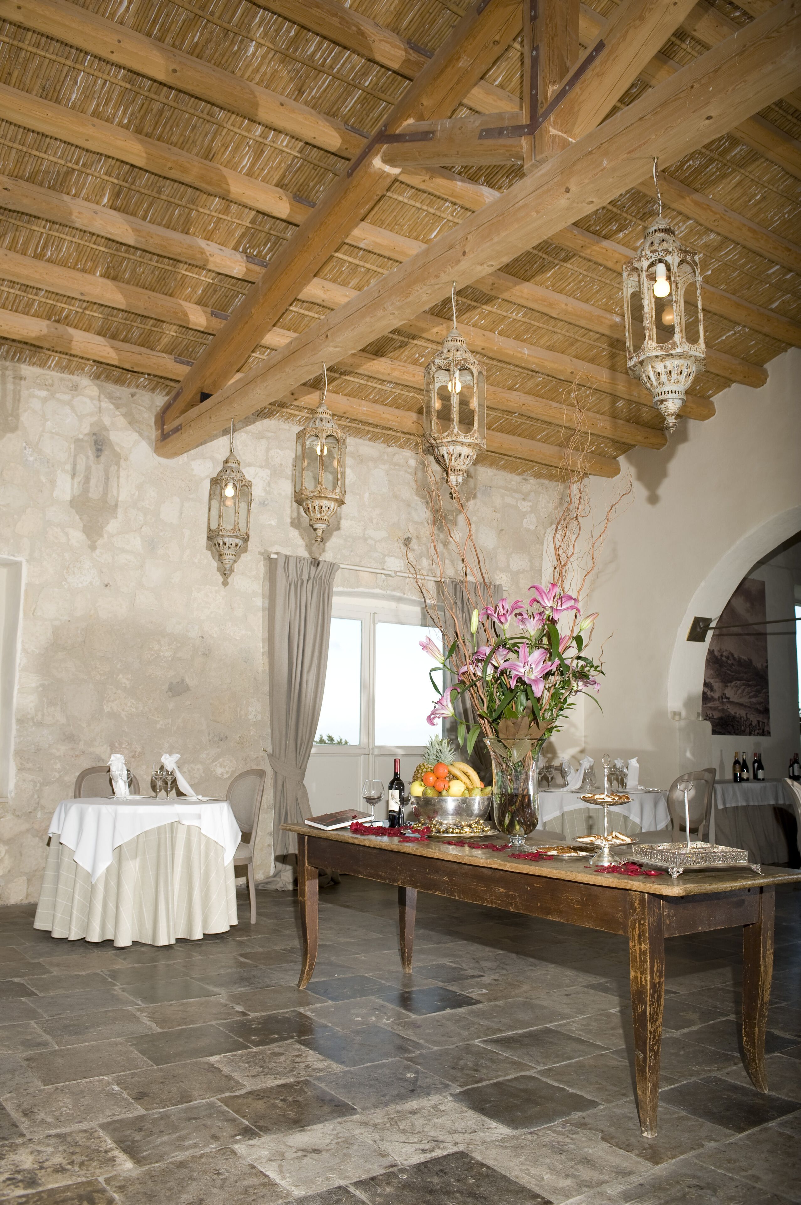 Un comedor rústico con una mesa de madera dispuesta para una comida, un jarrón de flores y faroles colgantes.