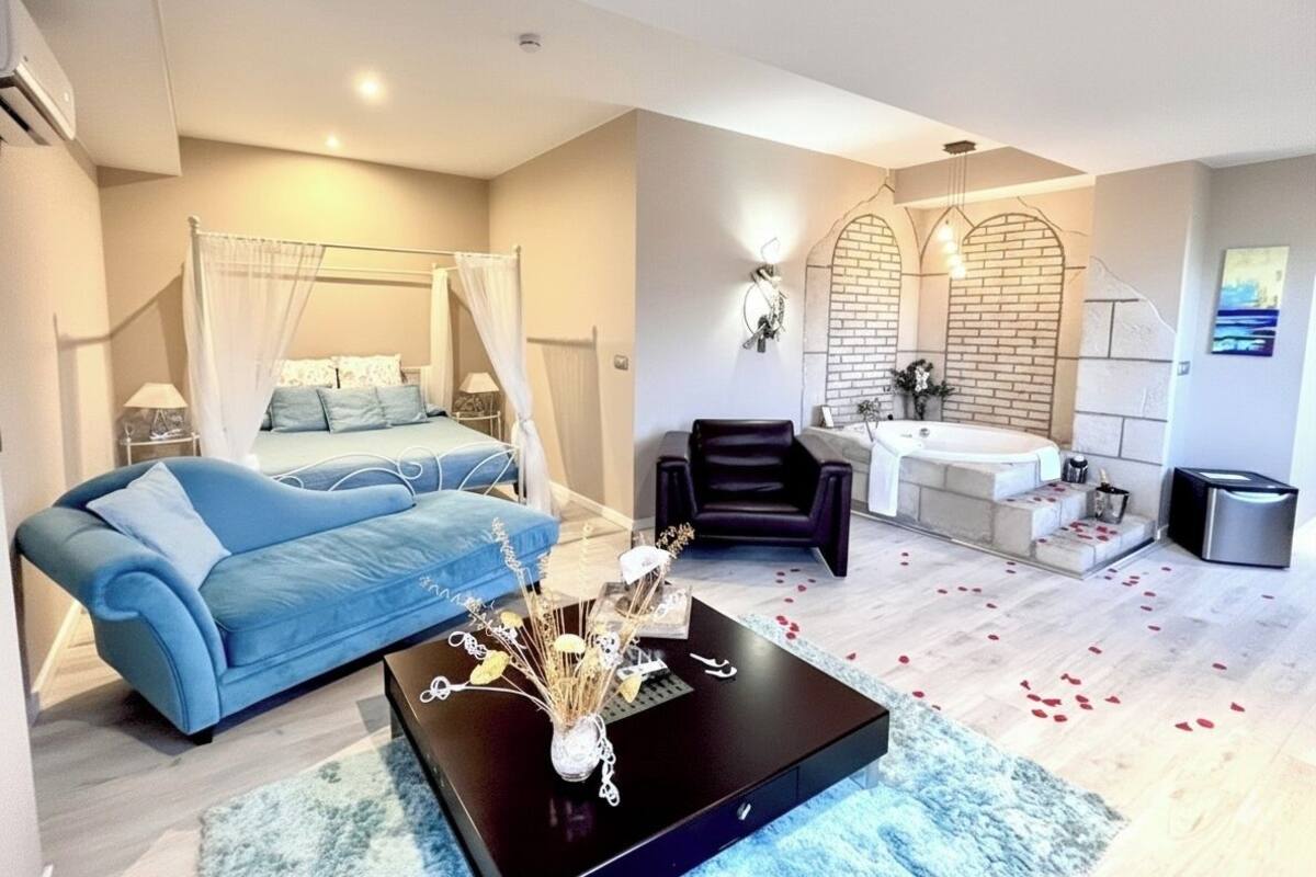 Une chambre à coucher moderne avec un lit, un canapé bleu, une table basse noire agrémentée d’objets décoratifs, un fauteuil en cuir noir, une baignoire blanche et un petit lavabo.