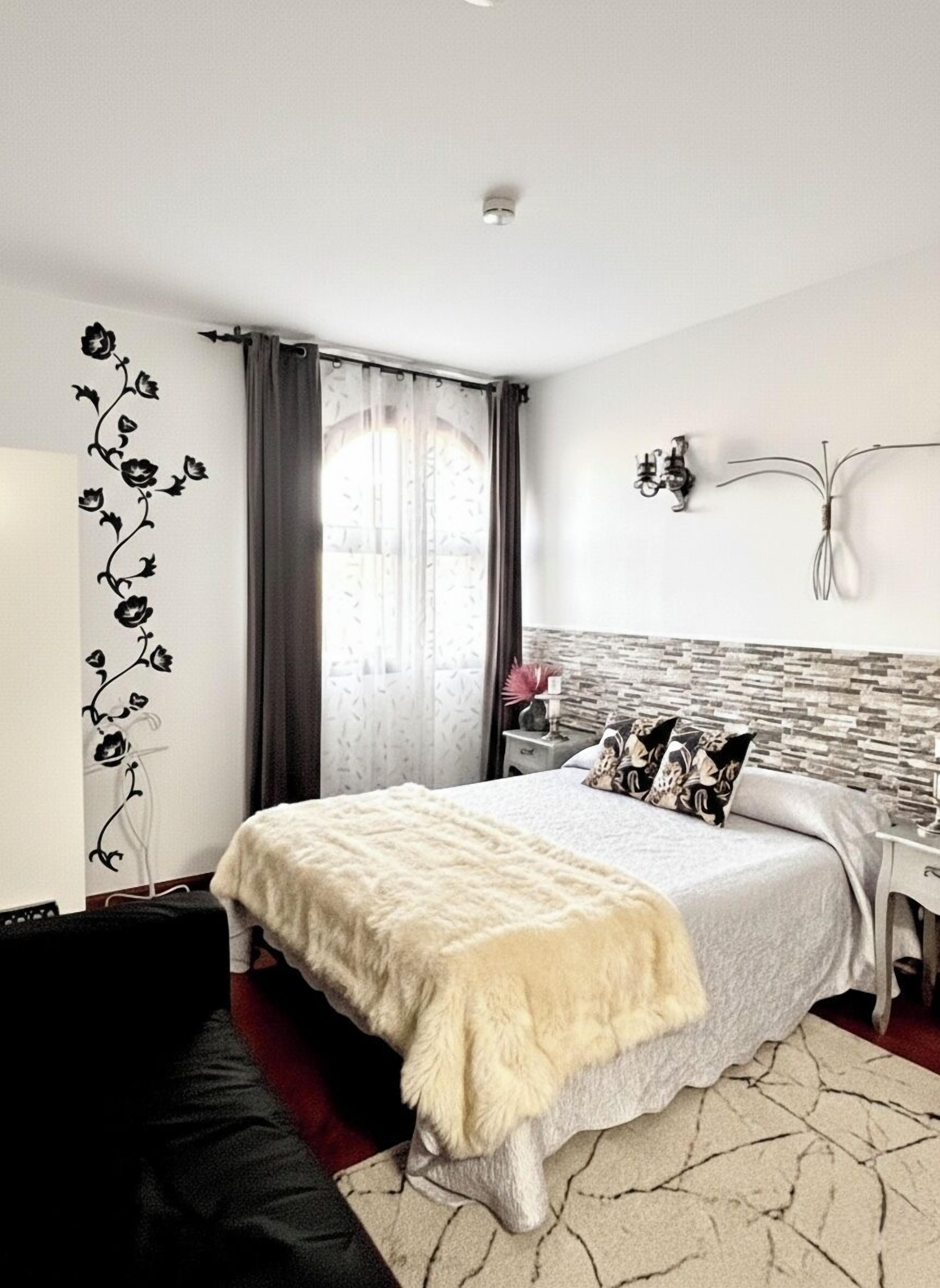 A bedroom with a bed, a nightstand, a lamp, a wall-mounted light fixture, and a window with curtains.