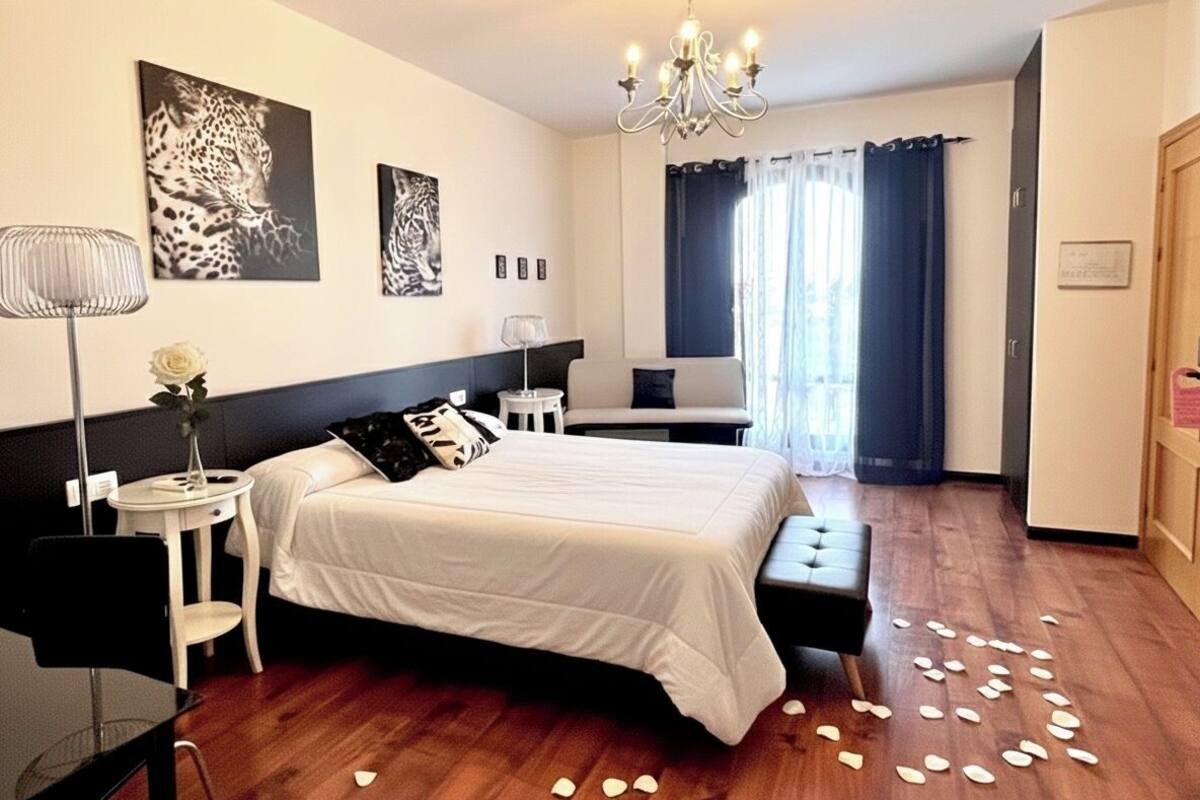 Une chambre à coucher comprenant un lit, des tables de chevet, un bureau, une chaise, un lustre et une fenêtre avec des rideaux.
