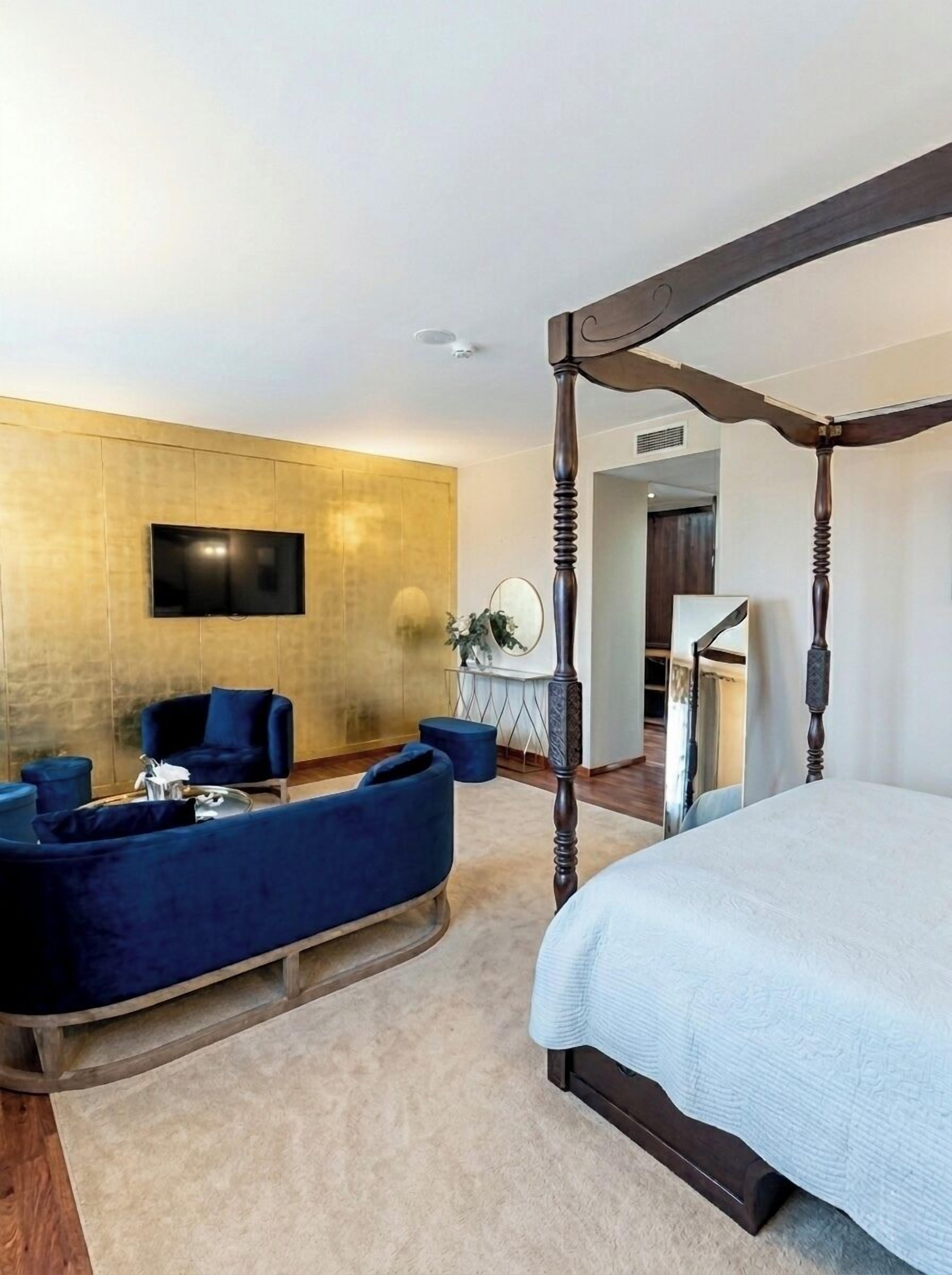 A bedroom with a four-poster bed, a TV mounted on the wall, a seating area with sofas, and a small table with a mirror.