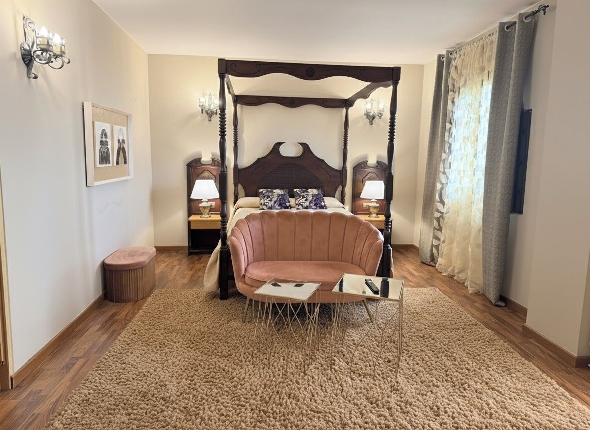 Un lit à baldaquin, une chaise longue, une petite table ronde, deux tables de chevet avec des lampes, un tableau encadré au mur et une fenêtre avec des rideaux.