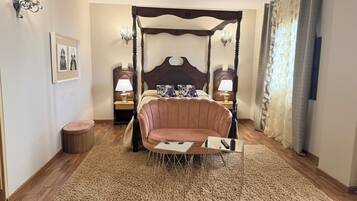 Una cama con dosel, un sillón de descanso, una mesita redonda pequeña, dos mesitas de noche con lámparas, un cuadro enmarcado en la pared y una ventana con cortinas.