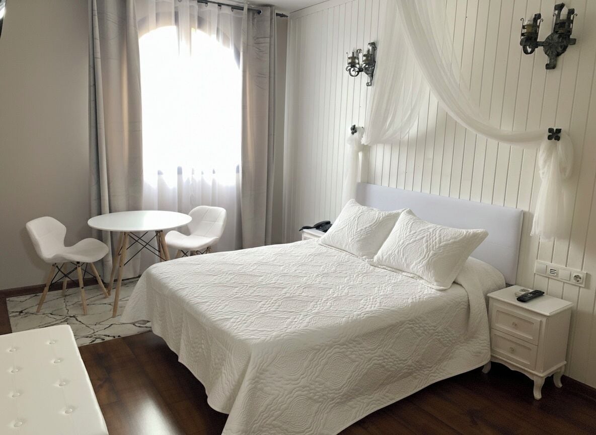 Una habitación de hotel con una cama grande, una mesita de noche, una silla, una pequeña mesa redonda y un ventanal con cortinas.