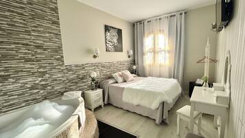 Un dormitorio moderno con una bañera grande, una cama con edredón blanco, una mesita de noche con un florero y un escritorio con espejo.