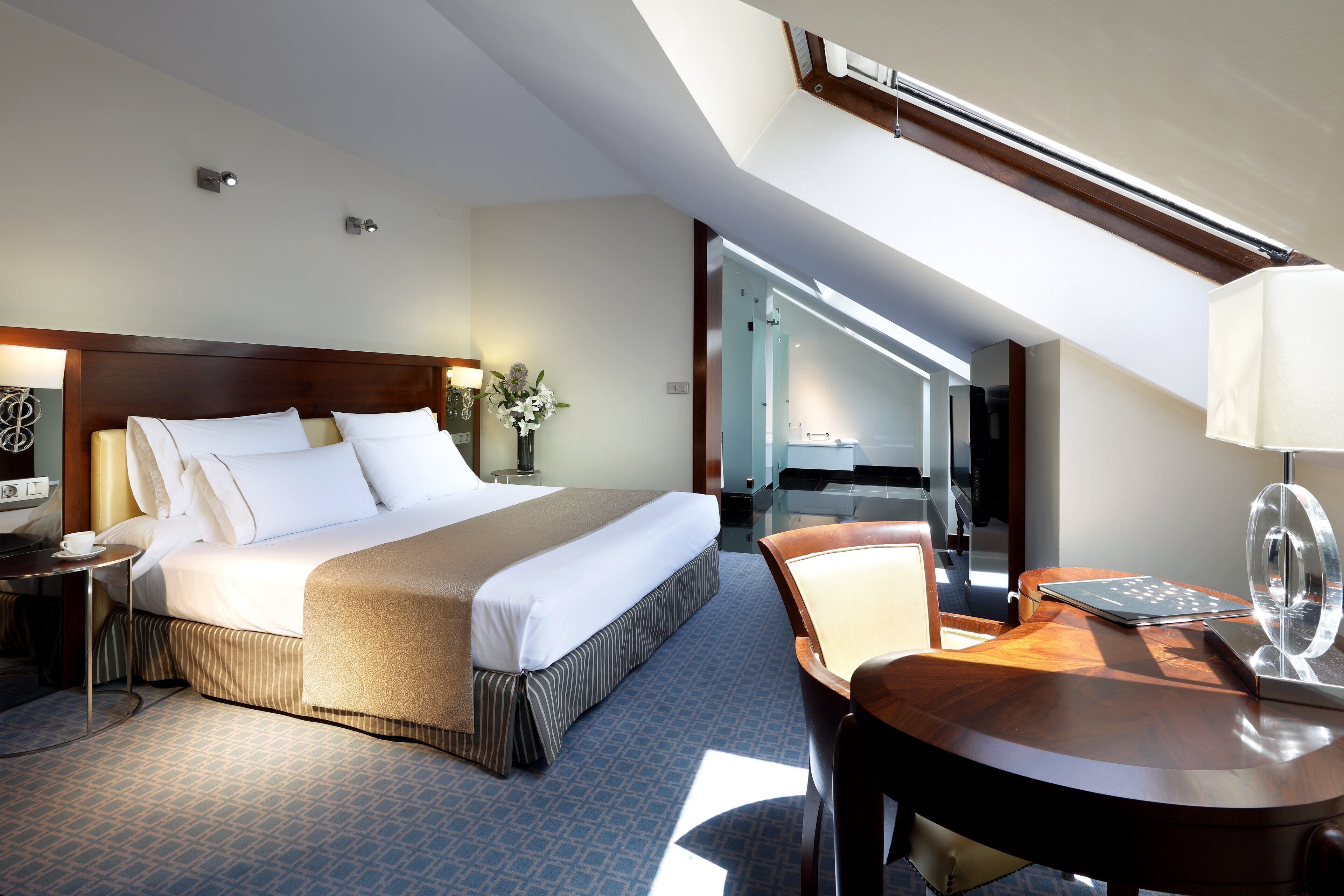 A hotel room with a large bed, a desk with a lamp, and a view of the hallway.