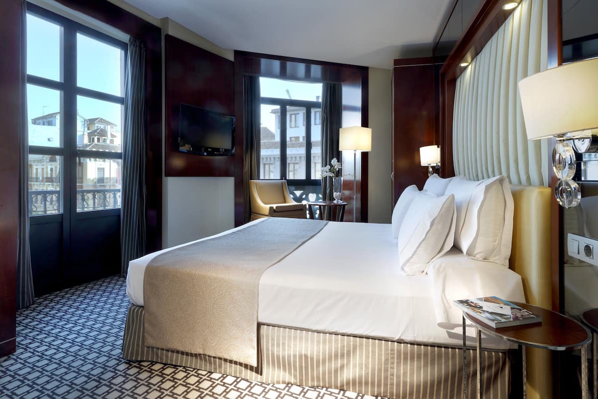 A hotel room with a large bed, a TV mounted on the wall, a window with curtains, and a view of buildings outside.