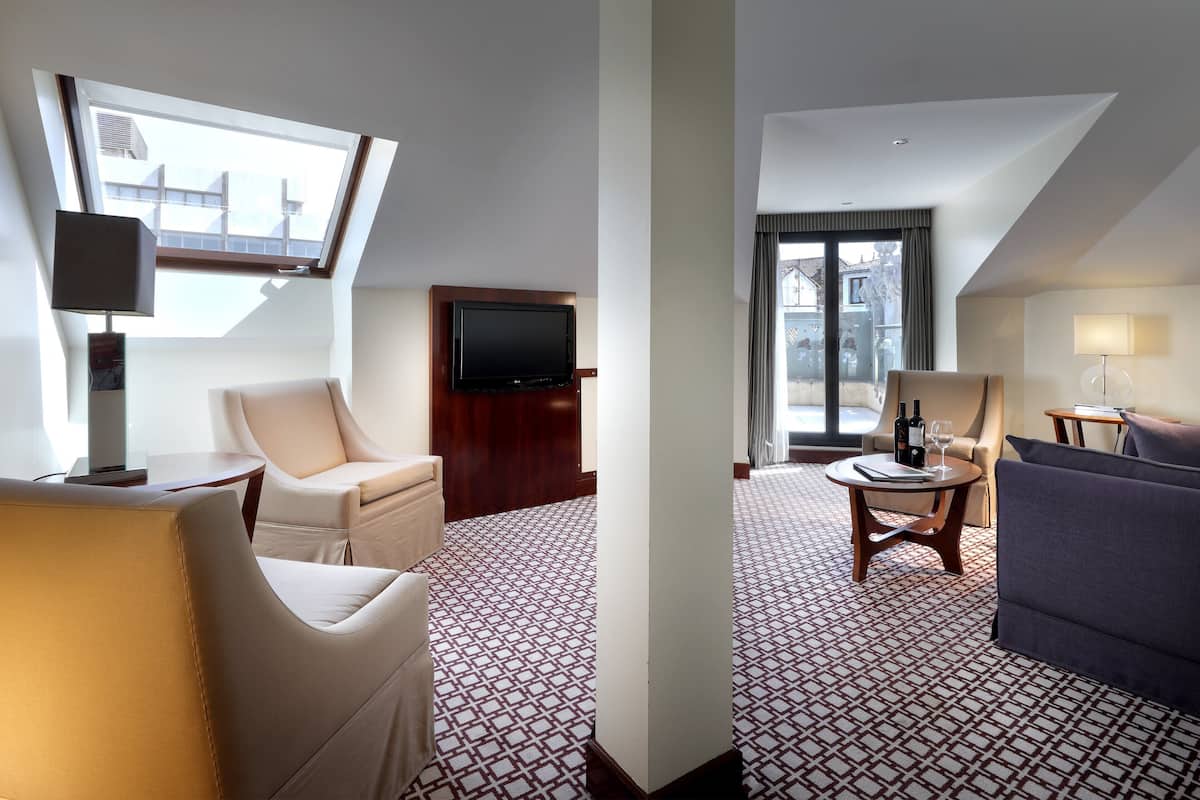 A hotel room with a large window, a flat-screen TV, a sofa, a chair, and a patterned carpet.