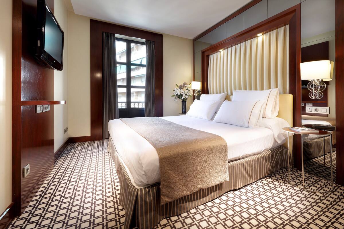 A hotel room with a large bed, a television mounted on the wall, a bedside table with a lamp, and a patterned floor.