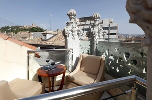 Un balcon avec une chaise beige, une petite table ronde sur laquelle se trouve un verre de vin rouge, et une vue sur un paysage urbain comprenant une colline surmontée d’une structure ressemblant à un château.