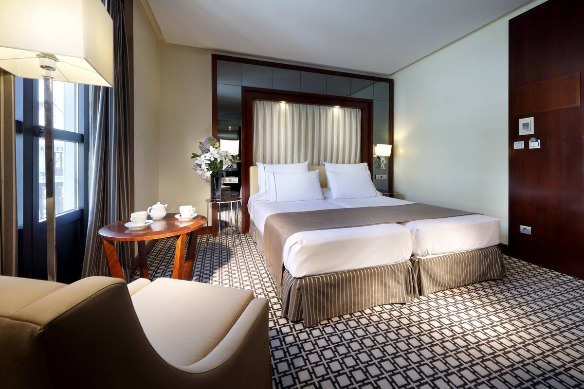 A hotel room with a large bed, a small table with a teapot and cups, a chair, and a desk with a lamp.