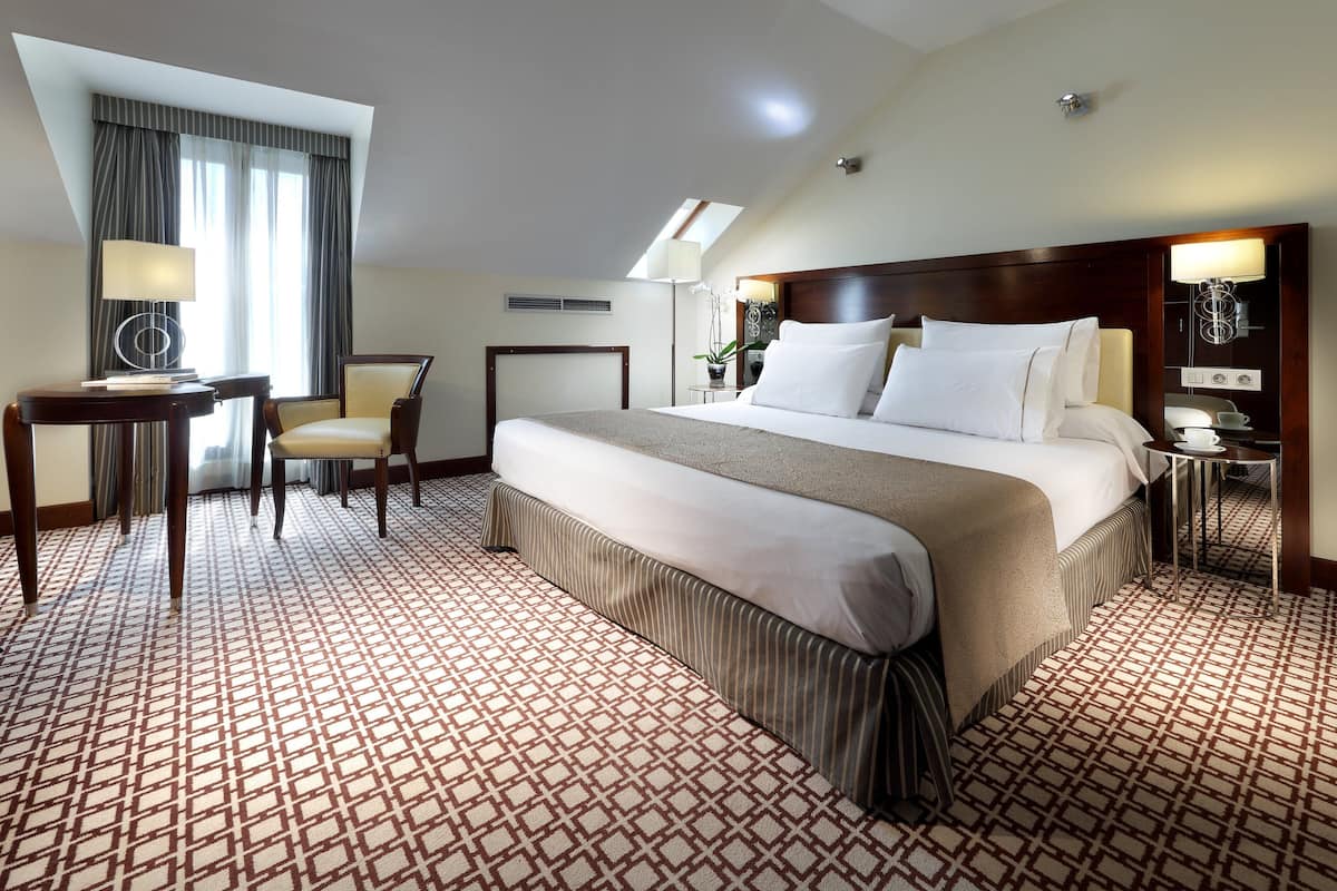A hotel room with a large bed, a desk with a lamp, a chair, and a small table with a plant.