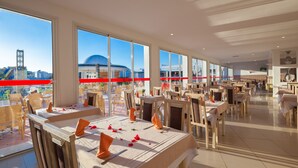 A spacious dining area with multiple tables and chairs, large windows offering a view of the cityscape, and a modern interior design. - Zodiac Hotel (Hammamet)