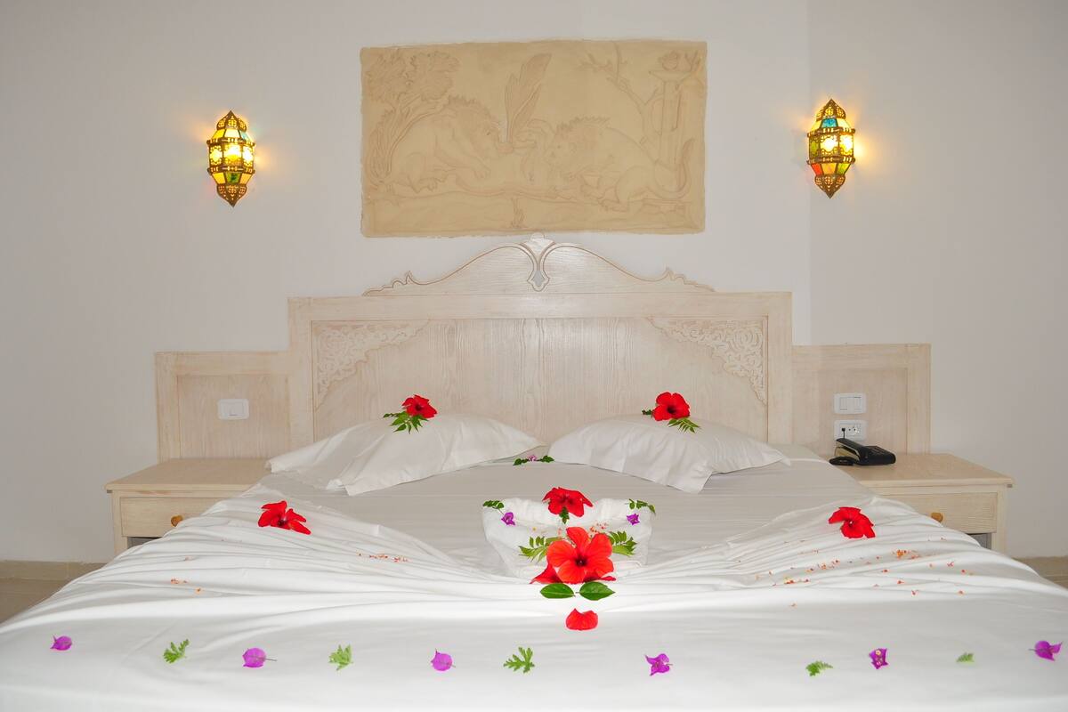 Una cama bien tendida con sábanas blancas, flores rojas y un tapiz decorativo en la pared.