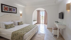 A hotel room with a bed, a desk, a TV, and a view through an arched doorway. - Zodiac Hotel (Hammamet)