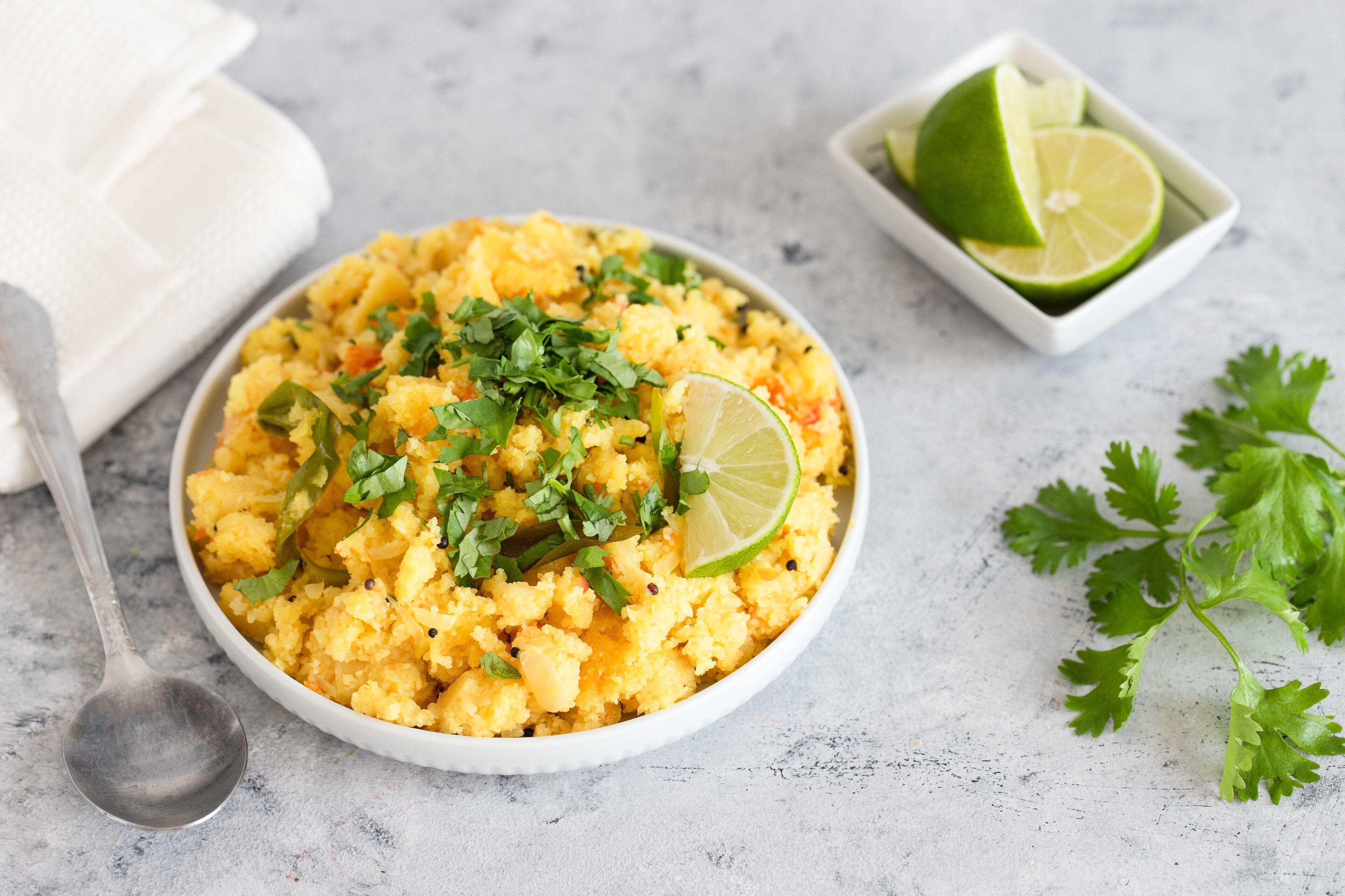 Un tazón de comida amarilla adornada con hierbas y lima, con una cuchara y un pequeño recipiente con rodajas de lima sobre una superficie texturizada.