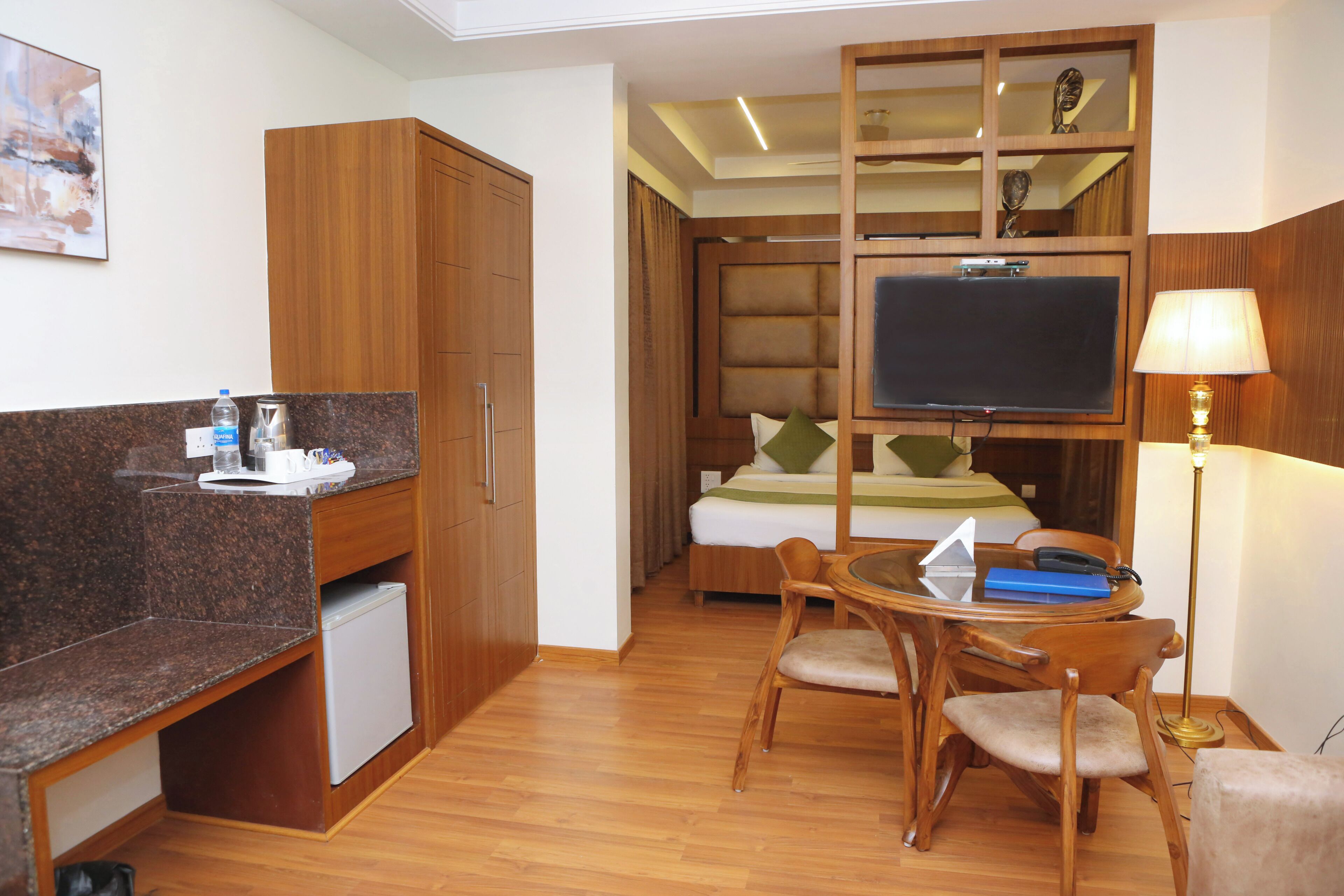 A hotel room with a kitchenette, a bed with a TV, a desk with a chair, and a lamp.