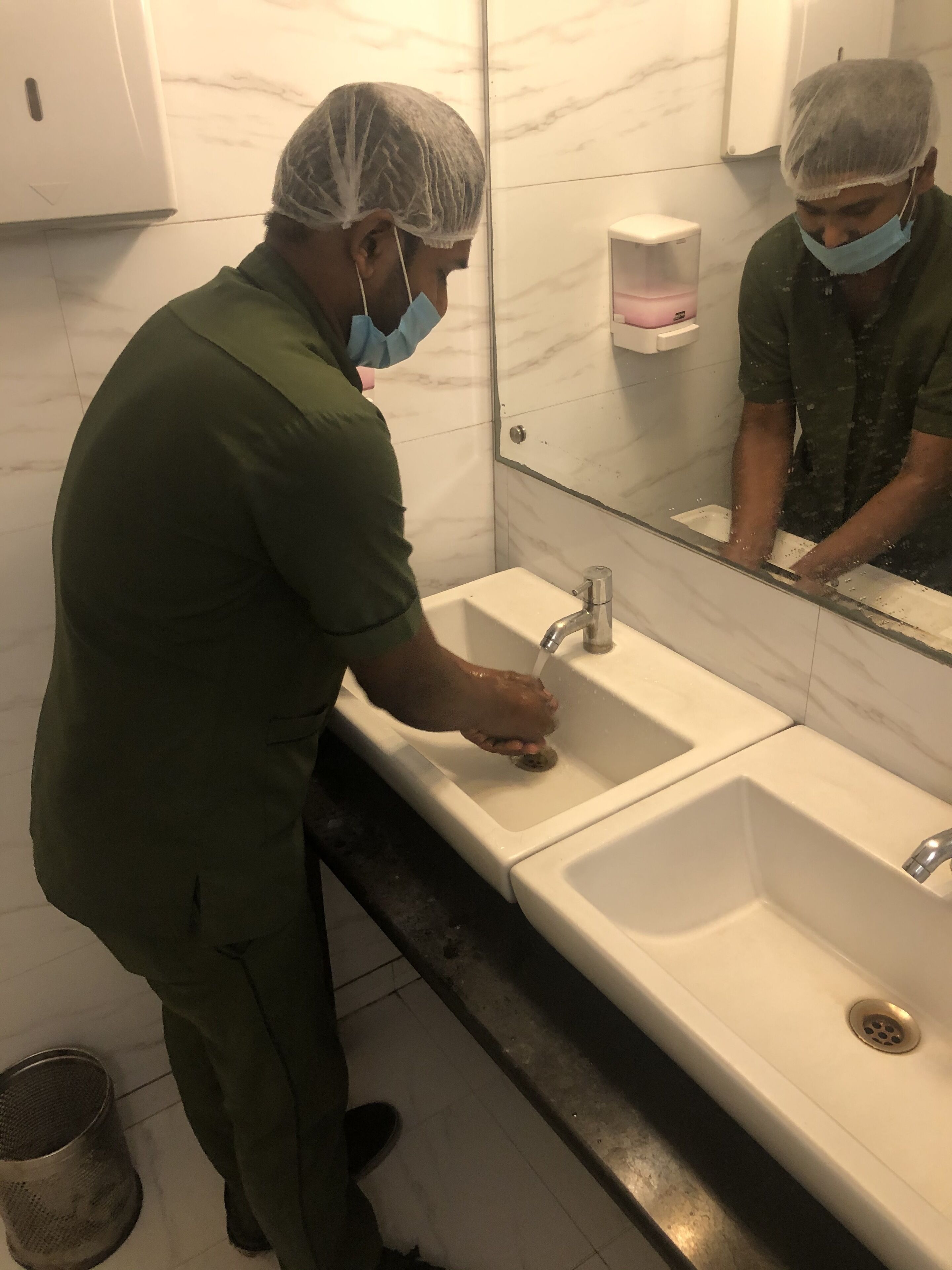 A person in a green uniform and hairnet washing hands in a bathroom with marble walls and a large mirror.