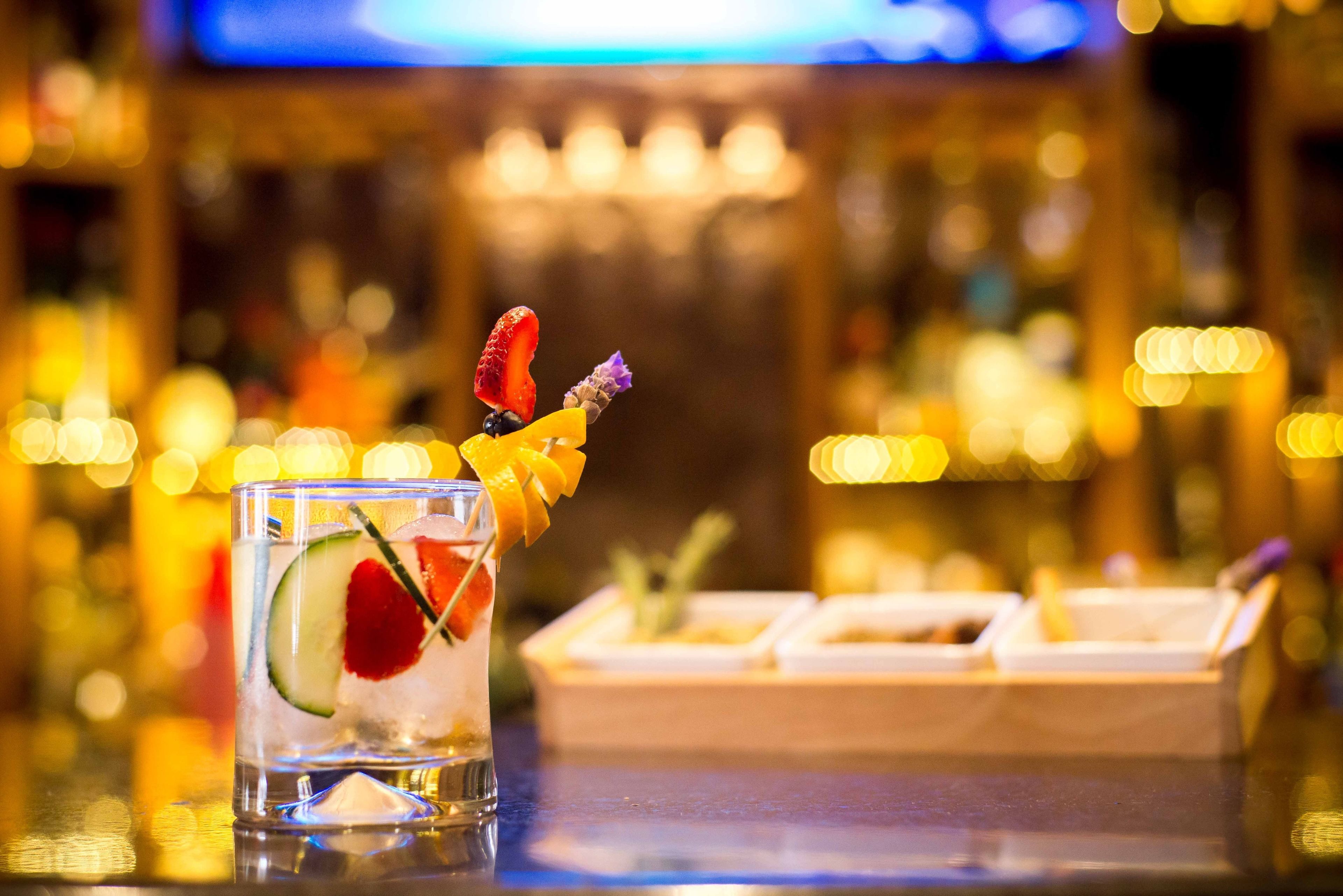 A cocktail with fruit garnish in a glass, a bar area with a blurred background, and a wooden bar counter.