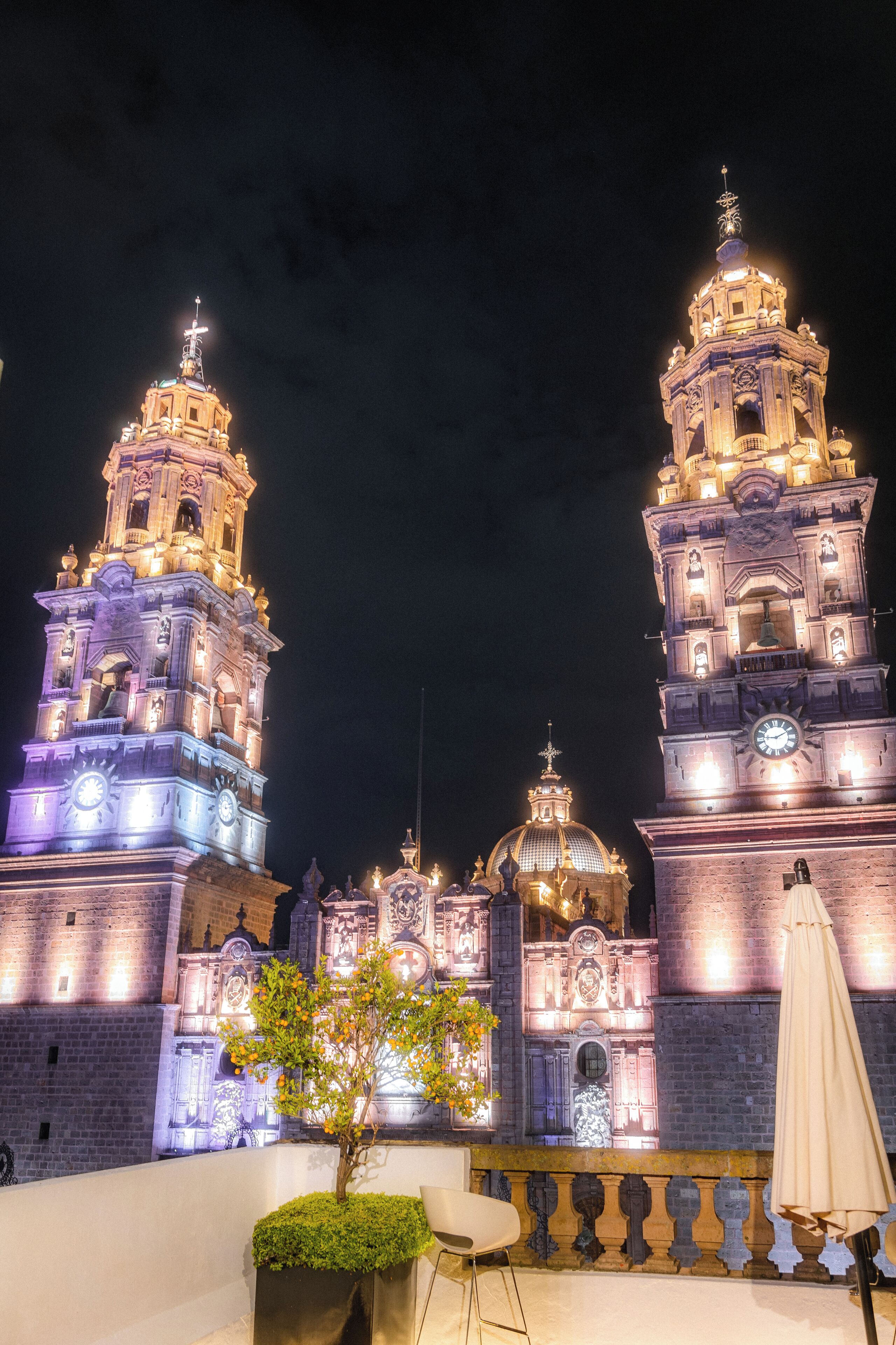 A historic building with illuminated towers and domes, a small tree, and outdoor seating.