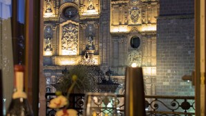 Una mesa de comedor con una botella de vino, una copa y un florero con flores. Al fondo, se observa un edificio de arquitectura ornamentada y una torre del reloj.