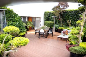 Een houten terras met een tafel en stoelen, omgeven door weelderige begroeiing en plantenbakken.
