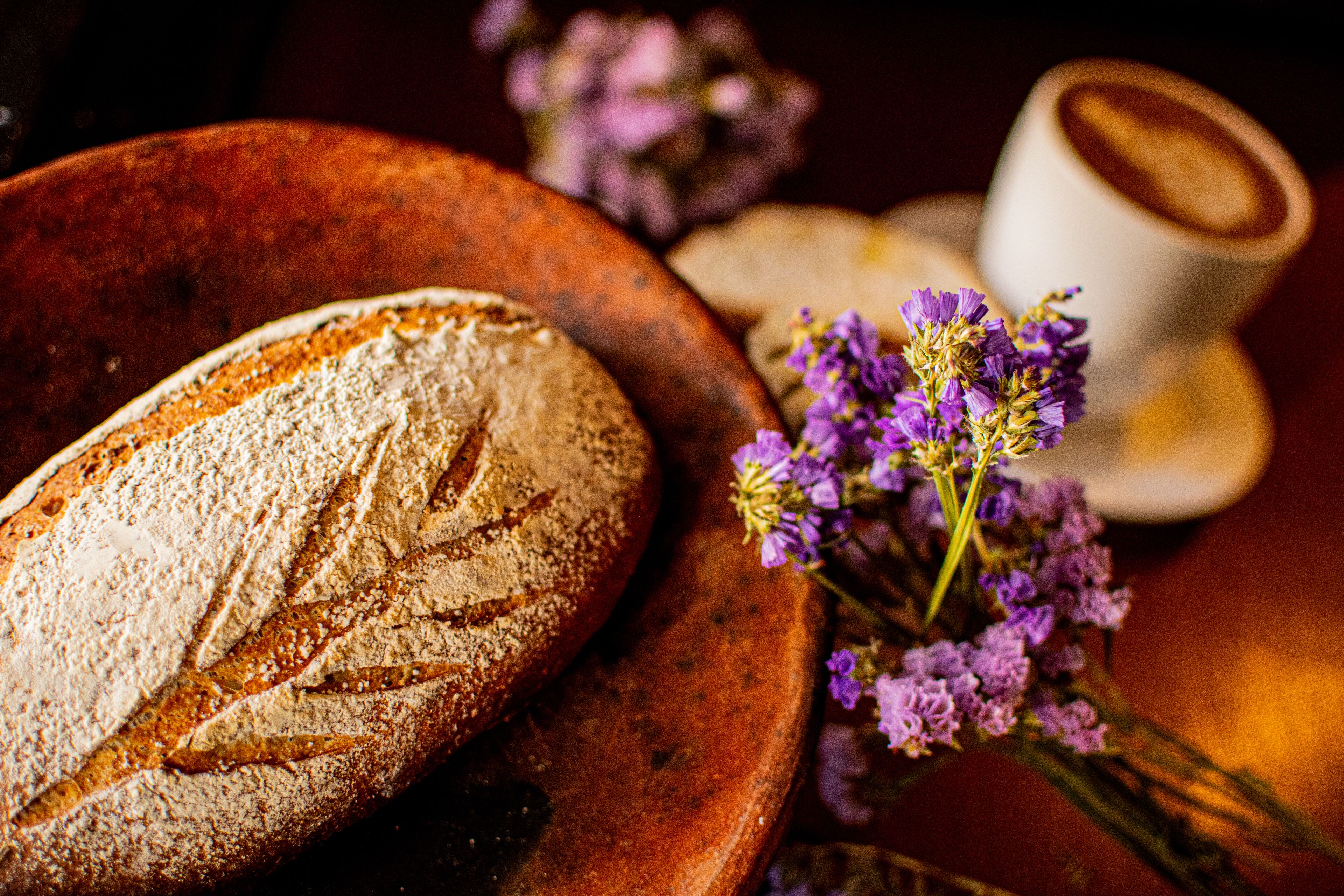 A loaf of bread on a wooden plate, a cup of coffee, and purple flowers.