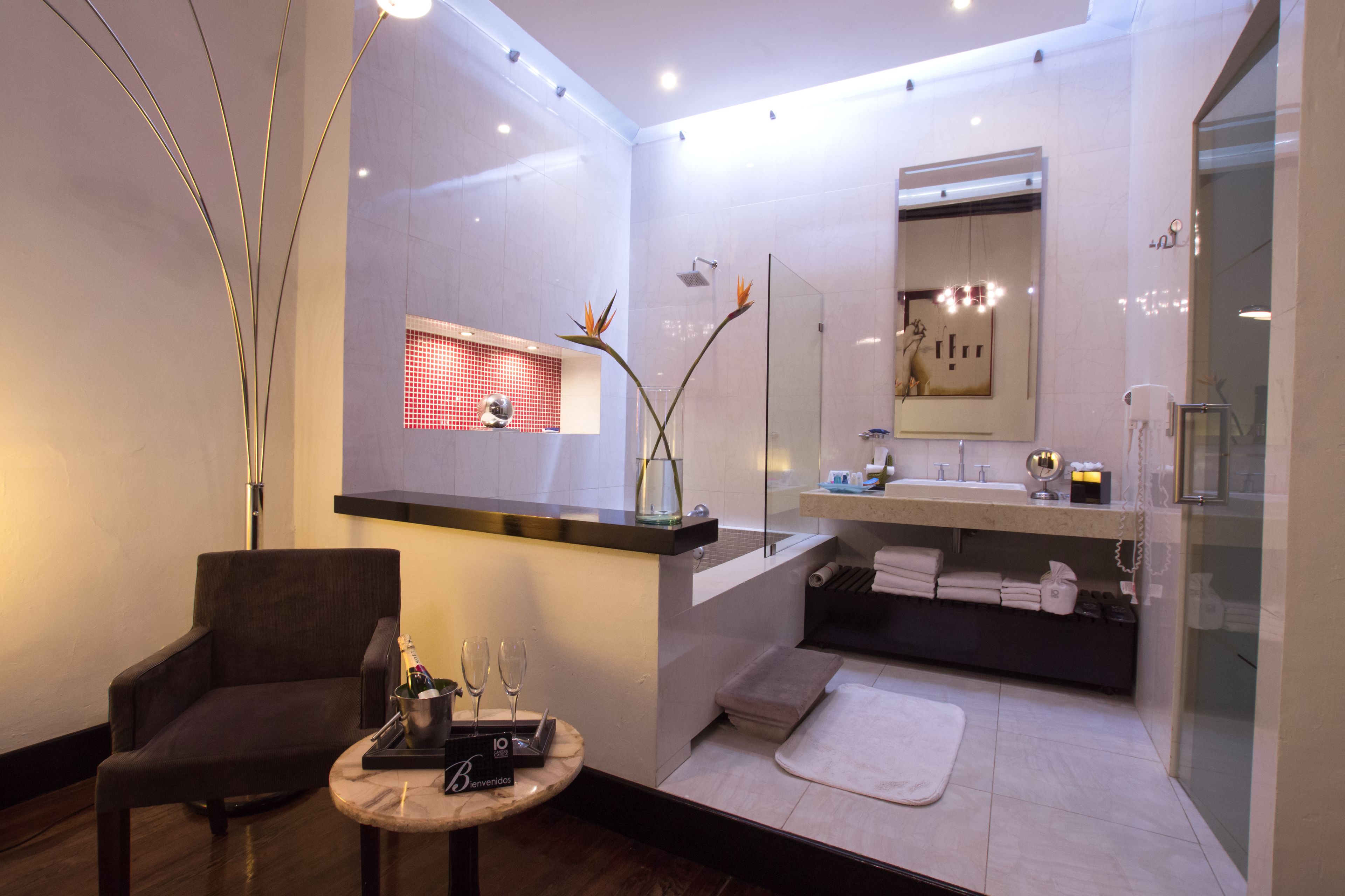 A modern spa room with a dark armchair, a small table with a bottle and glasses, a glass shower enclosure, a large mirror, and a white tiled floor.