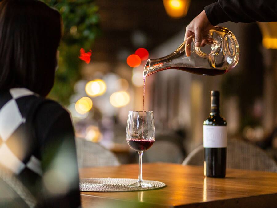 A person pouring wine into a glass on a wooden table with a bottle of wine nearby.