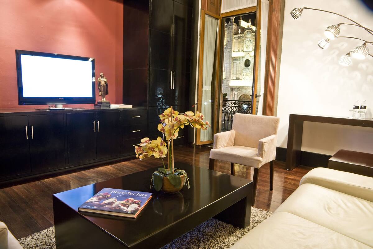 Una sala moderna con una televisión de pantalla plana, una mesa de centro con un libro y flores, un sillón blanco y un mueble de madera oscura.