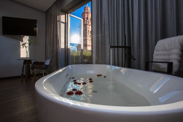 Quarto de hotel moderno com uma banheira grande, uma TV presa na parede e vista para uma torre histórica pela janela.