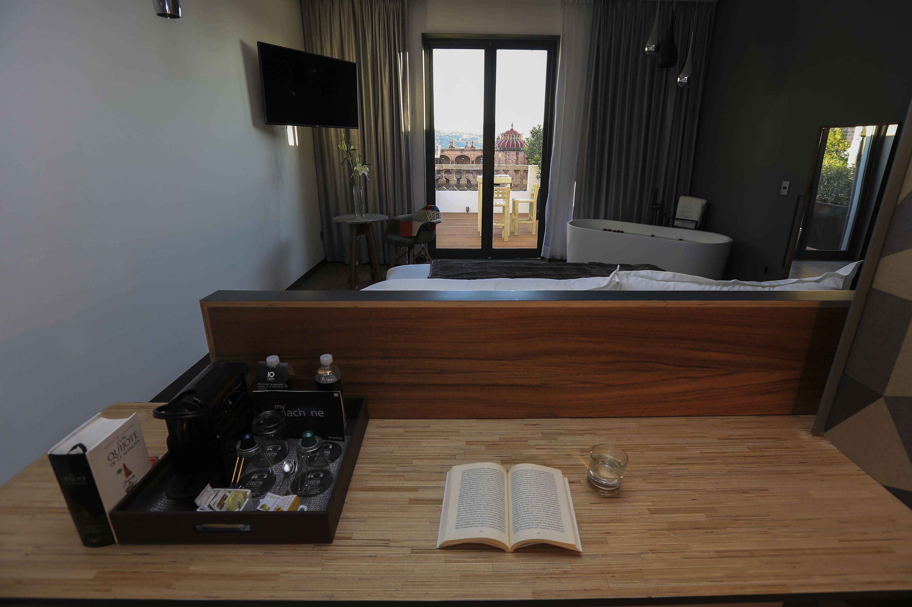 Habitación de hotel con cama, una mesa de madera con un libro y una cafetera, televisión y balcón con vistas a edificios.