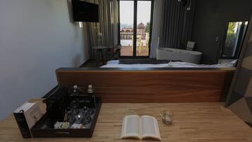 Habitación de hotel con cama, una mesa de madera con un libro y una cafetera, televisión y balcón con vistas a edificios.