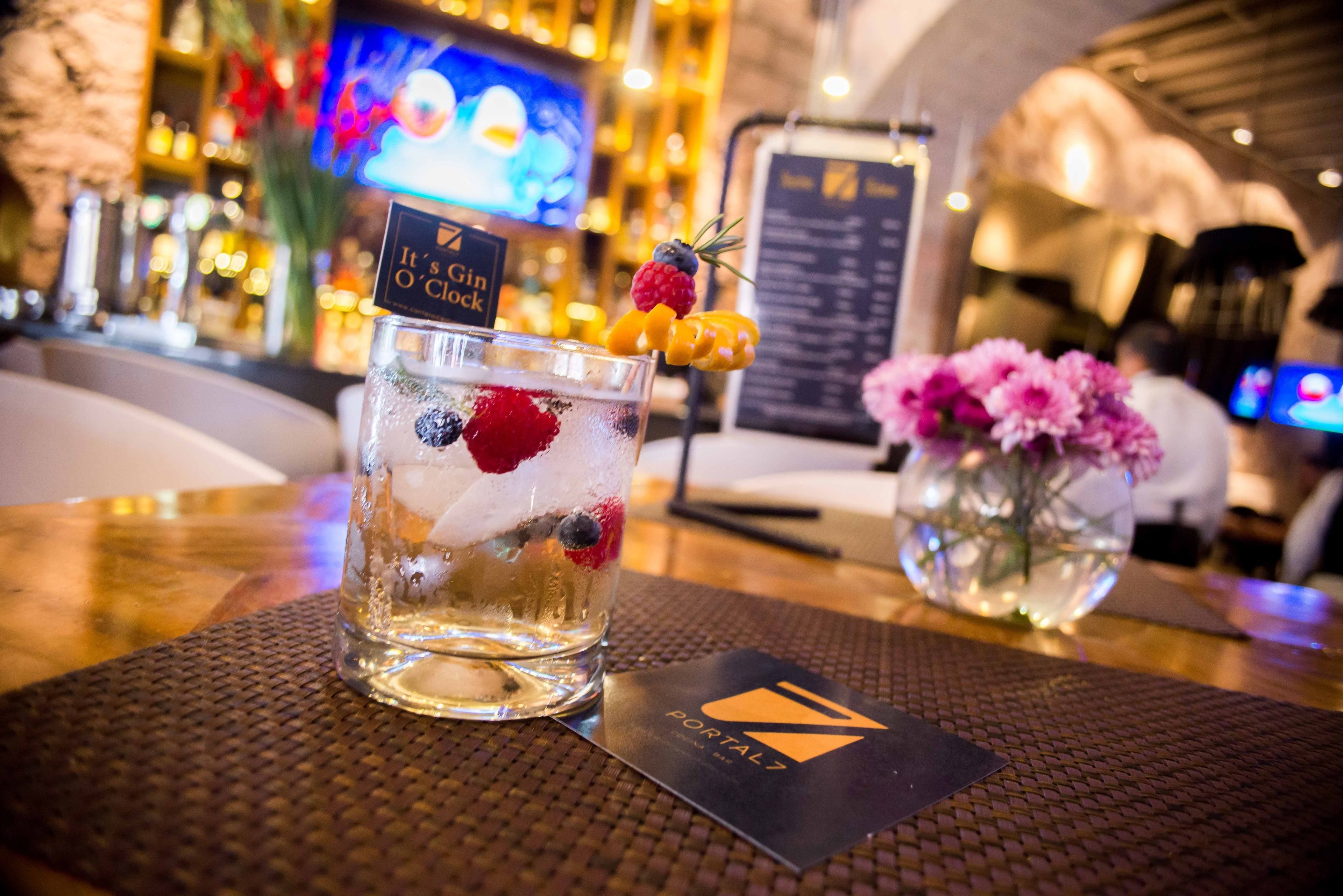 A bar setting with a cocktail glass garnished with berries and a sprig of rosemary, a menu board, and a vase with pink flowers on a table.