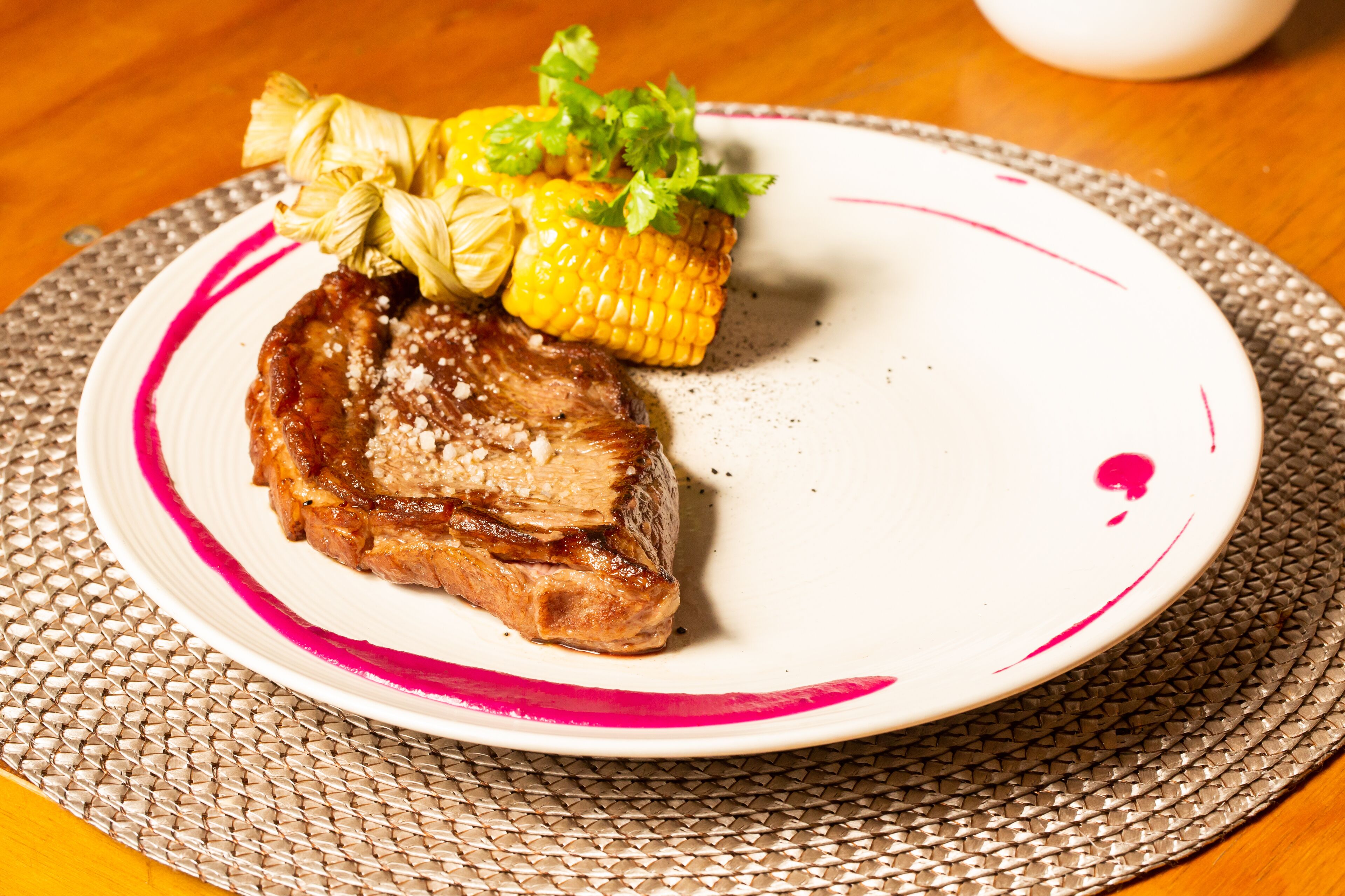 A plate with a piece of meat, corn on the cob, and a garnish on a woven placemat.