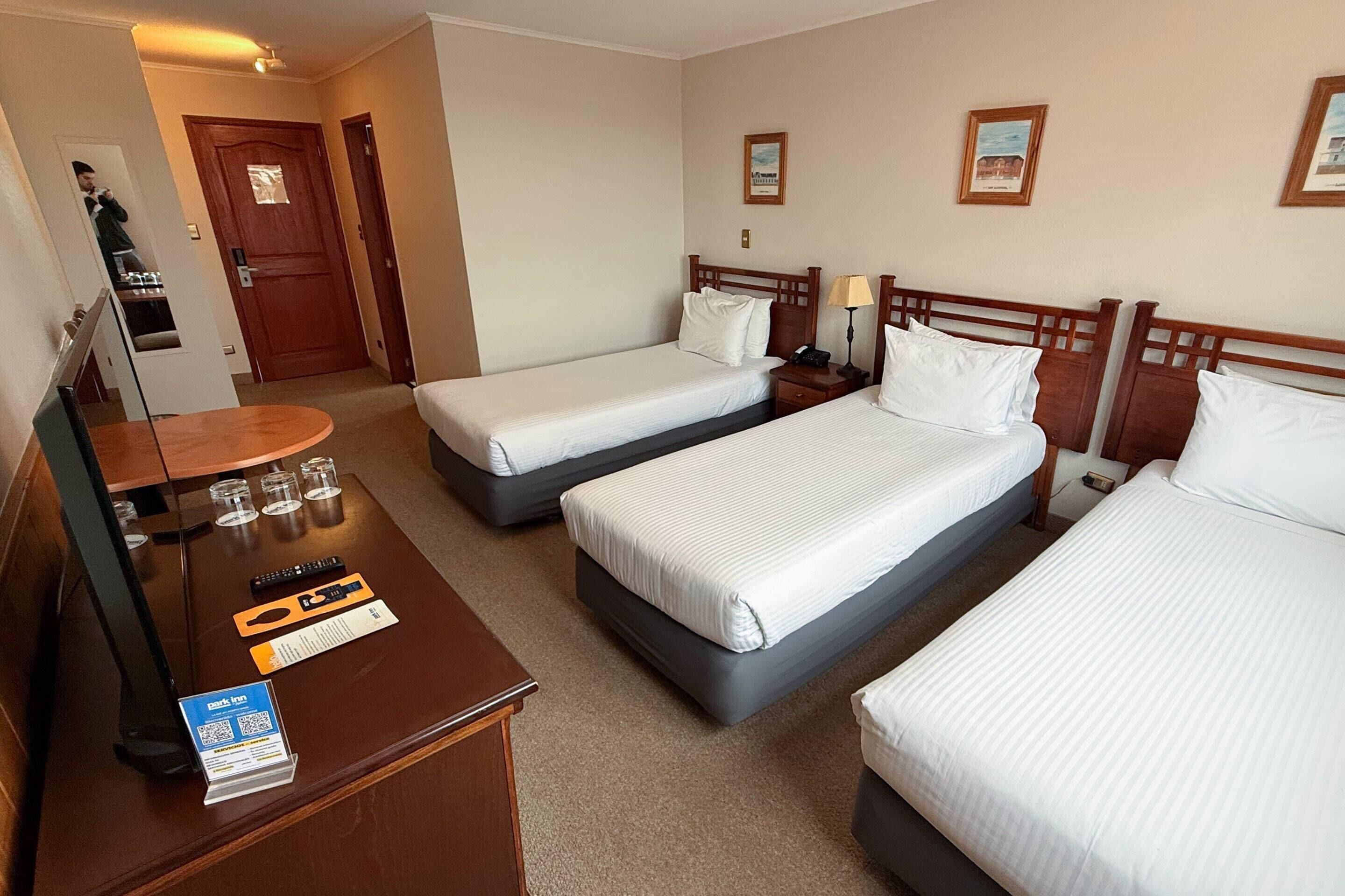 A hotel room with two single beds, a wooden desk with a mirror, a small table with glasses, and a framed picture on the wall.