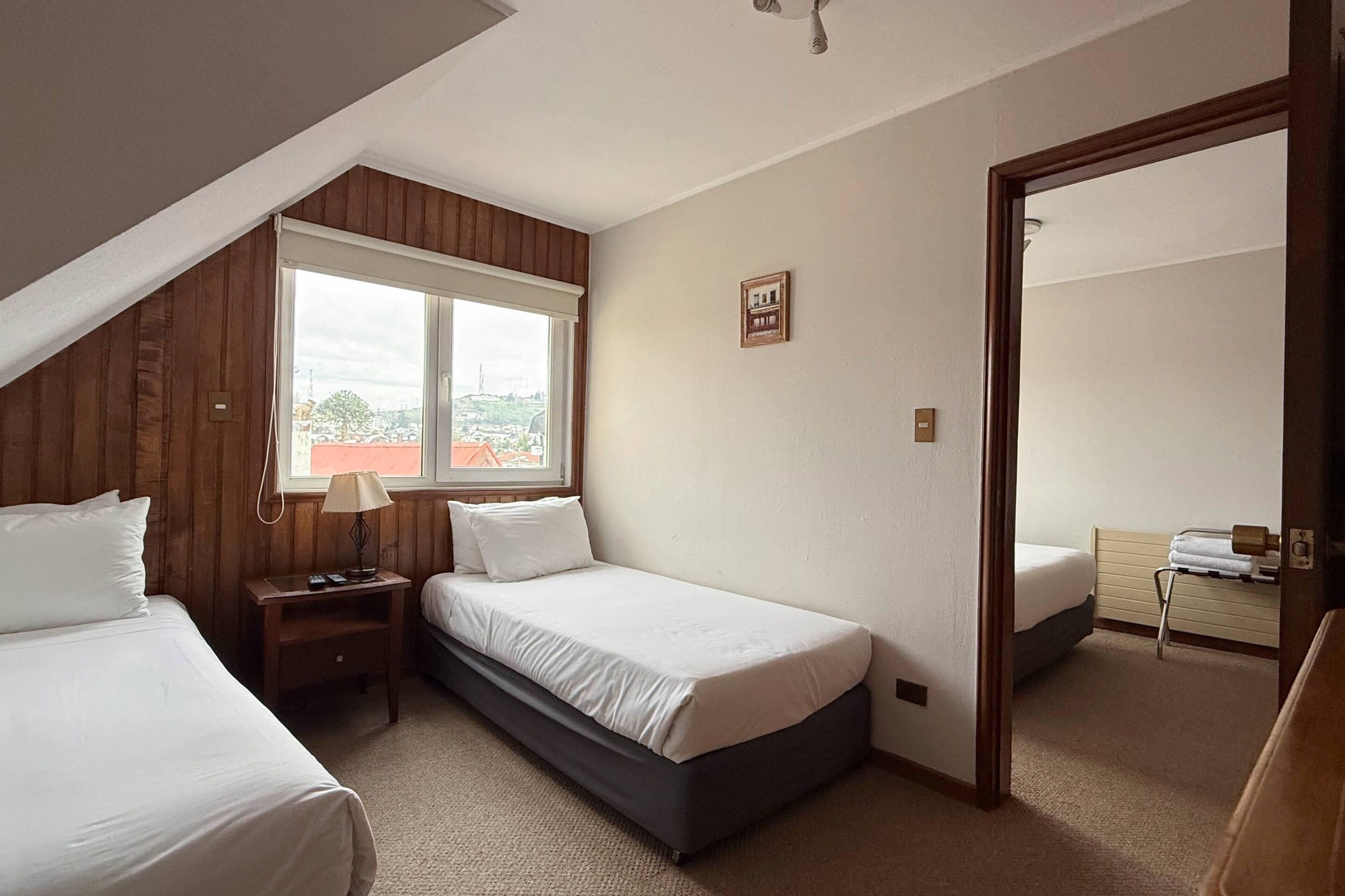 A bedroom with two beds, a window, a wooden wall, a small bedside table, and a mirror.
