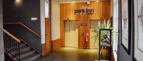 Un hall de hotel con una escalera, un mostrador de recepción, un cartel de 'Park Inn' y una planta en maceta.
