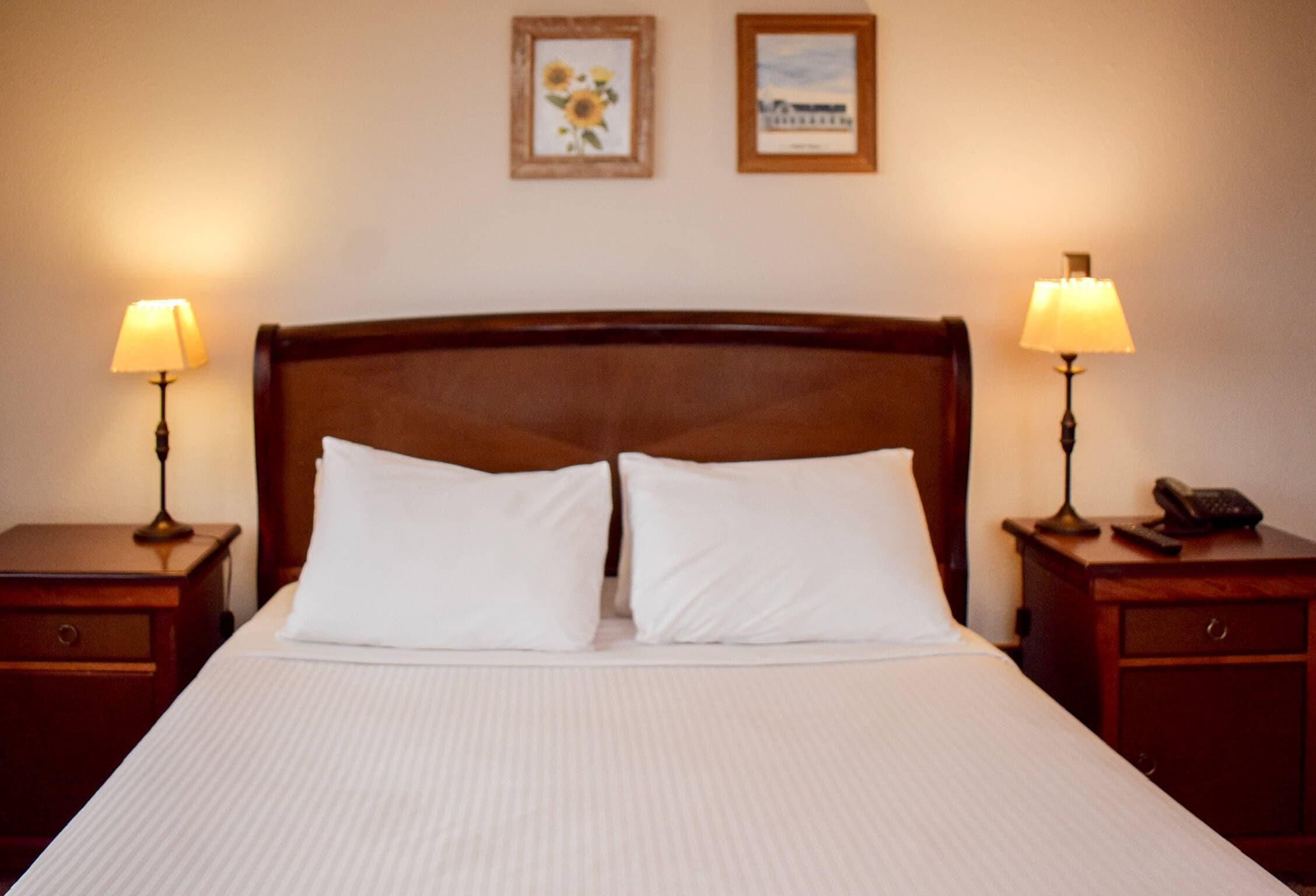 A neatly made bed with white linens, two bedside lamps, a wooden headboard, and two framed pictures on the wall.
