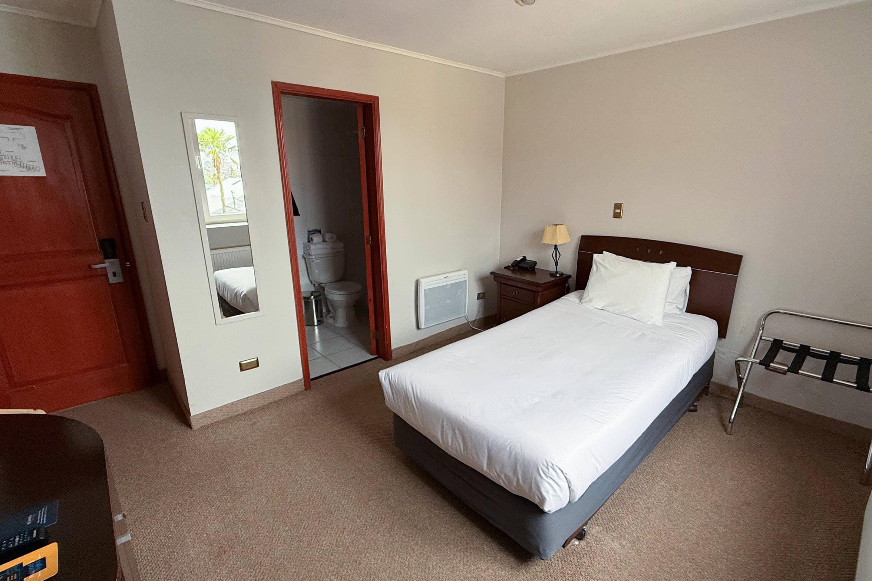 Habitación de hotel con cama, escritorio, silla, lámpara, radiador y puerta que da a un baño.