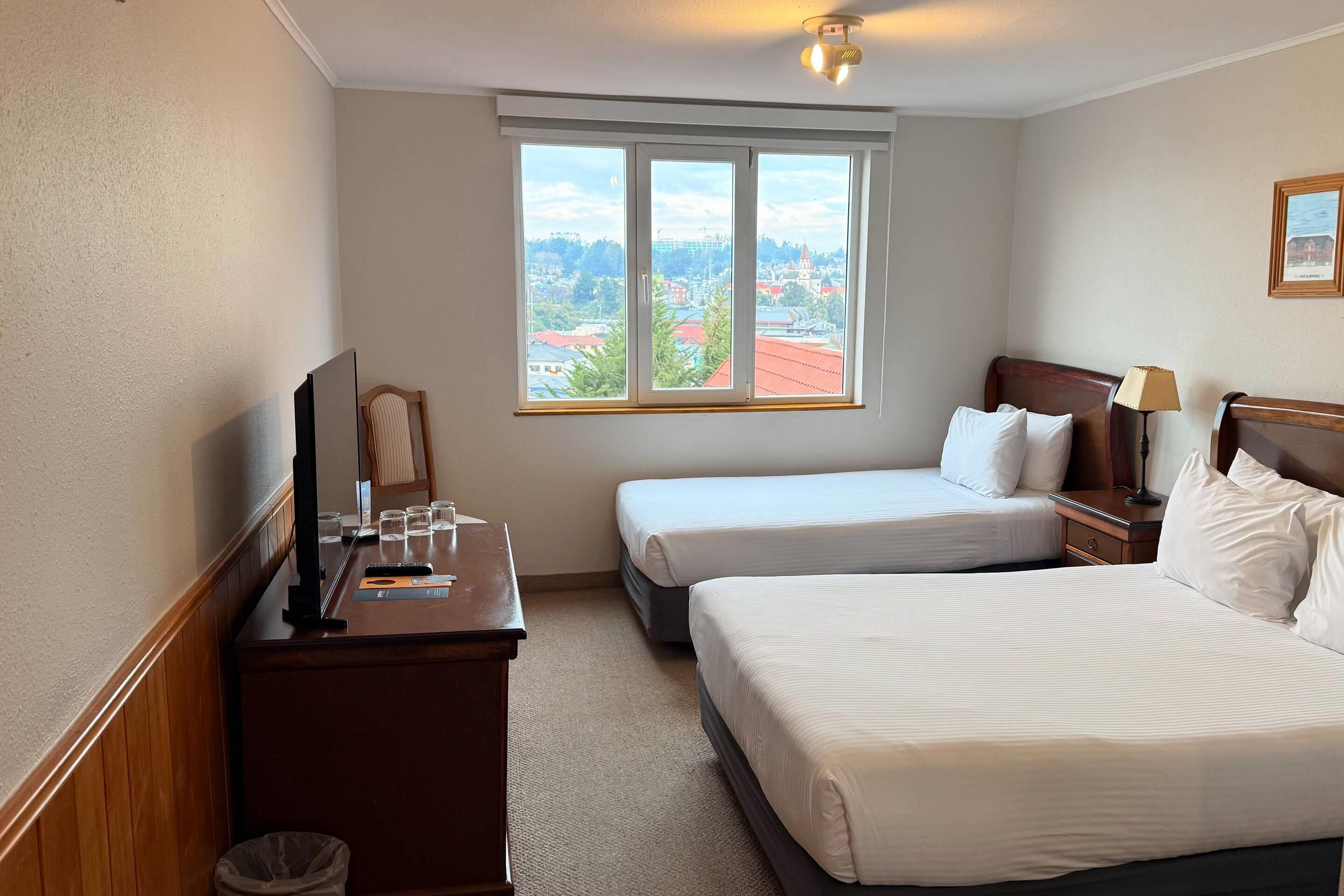 Une chambre d’hôtel avec deux lits, un bureau avec une télévision, une lampe et une fenêtre donnant sur la ville.