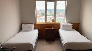 A room with two beds, a window with a view of a cityscape, a small table with a lamp, and a chair.