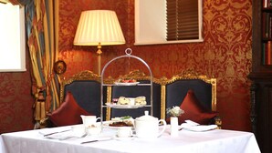 A three-tiered tea service stand on a table with white tablecloth, surrounded by red velvet chairs, a lamp, and a patterned wallpaper background.
