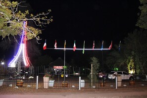 Une vue nocturne d’une zone délimitée par une clôture, avec une structure imposante et illuminée qui ressemble à la Tour Eiffel, une rangée de drapeaux et un bâtiment avec une enseigne en arrière-plan.