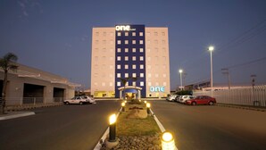 A hotel with a prominent 'one' sign, a parking lot with several cars, and a well-lit entrance pathway. - One Aguascalientes Sur (Aguascalientes)