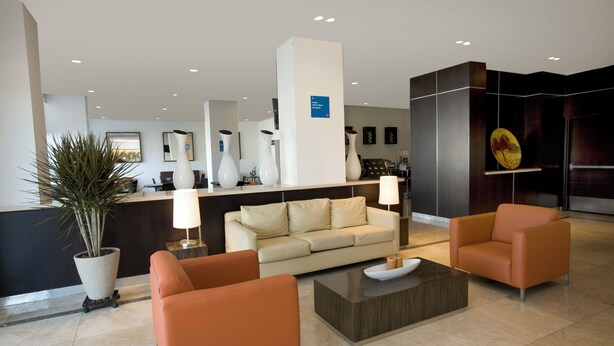 A modern lobby with a reception desk, a beige sofa, two orange armchairs, a coffee table, and a potted plant. - One Aguascalientes Sur (Aguascalientes)