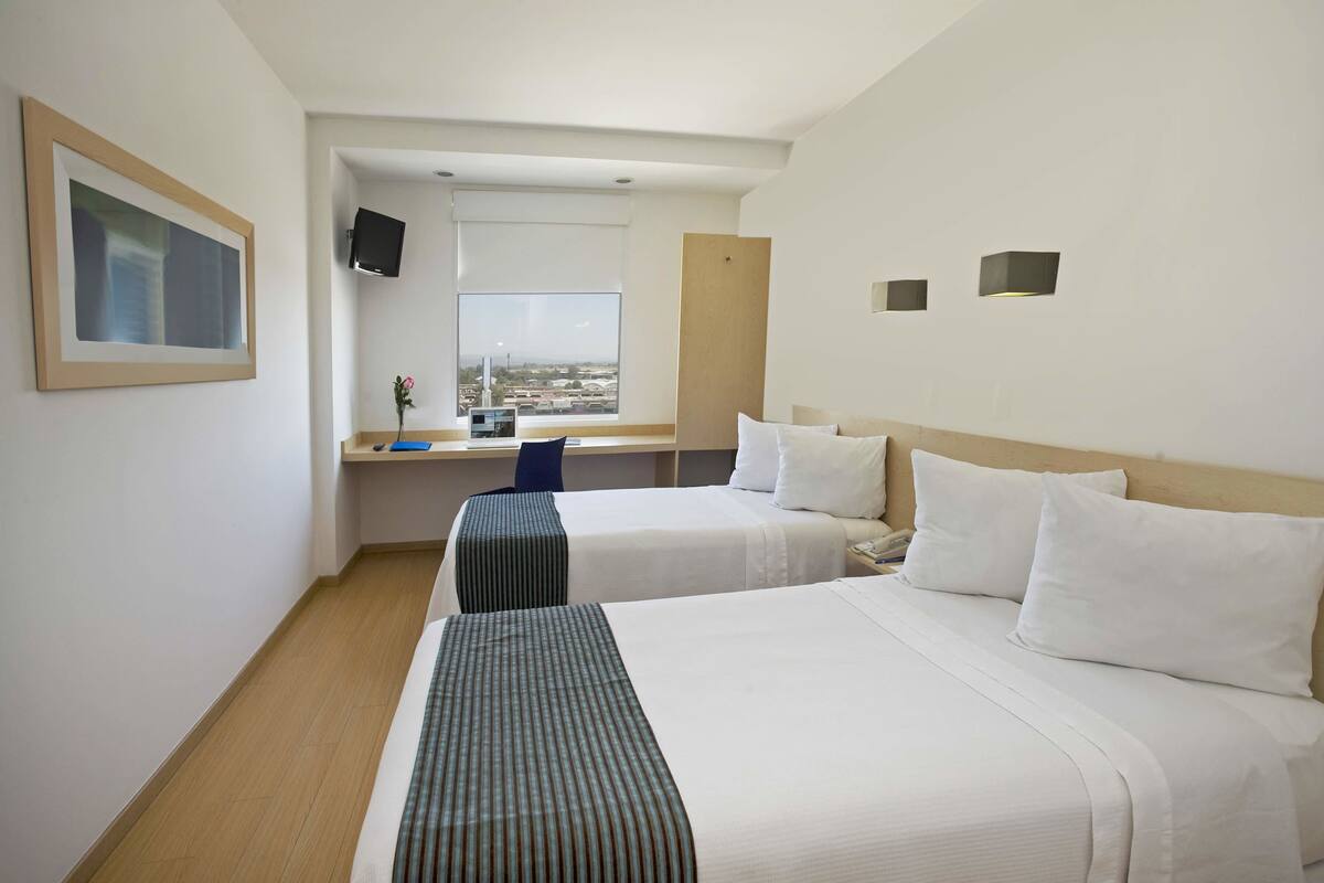 Habitació d'hotel amb dos llits, escriptori, televisió i finestra amb vista a la ciutat.